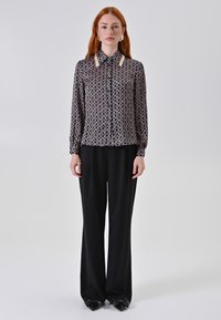 Donna con lunghi capelli rossi che indossa una blusa a fantasia bianco e nera, pantaloni neri a gamba larga e scarpe nere a punta con fiocchi.