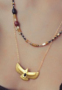 Pendentif en or représentant un aigle avec une pierre précieuse noire au centre. Accompagné d'une chaîne en perles avec des accents dorés, noirs et rouges.