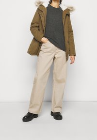 Olive green parka with a fur-lined hood, gray sweater, beige wide-leg trousers, and black ankle boots, styled in a casual outfit.