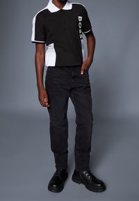 Polo nera con dettagli bianchi e logo, abbinata a jeans neri e stivaletti neri con lacci. L'outfit presenta un colletto classico e tre bottoni.