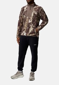 Tarnzip-Jacke mit einem Braun- und Beigemuster, kombiniert mit schwarzen Fleece-Hosen und grauen Sneakers mit weißen Sohlen.