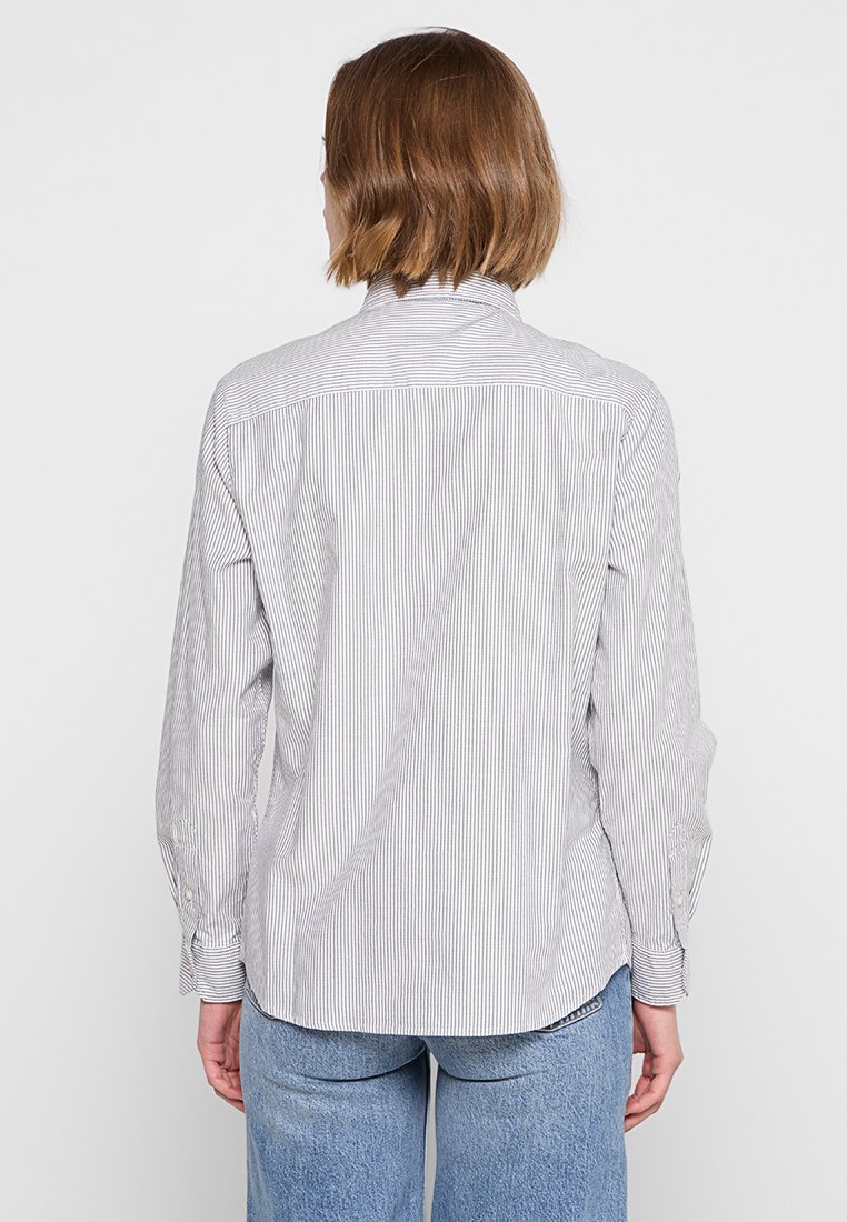 Persona con capelli corti castani che indossa una camicia bianca a maniche lunghe con sottili righe verticali e jeans blu, mostrata di spalle.