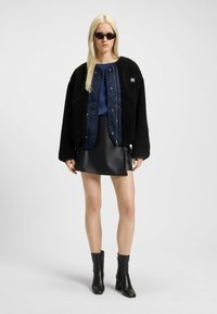Black textured jacket with a quilted inner lining, paired with a navy blouse and a black faux leather skirt. Model also wears black heeled boots.