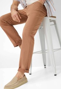Tan chinos met een slanke pasvorm, opgerolde zomen, gemaakt van katoen, gecombineerd met beige instappers op een metalen barkruk.