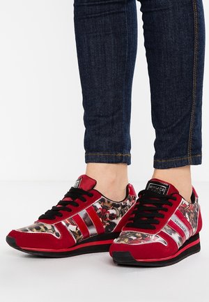 Czerwono-czarne sneakersy w wzory z czarnymi sznurówkami, noszone z ciemnoniebieskimi podwiniętymi dżinsami na białej podłodze.