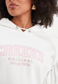 Sudadera blanca con capucha y cordón, con texto rosa "CREEKS ORIGINAL Desde 1974". Tejido suave y corte holgado. Pendiente texturizado dorado visible.
