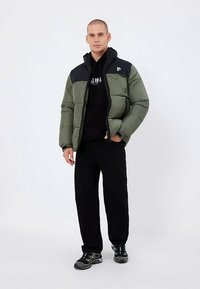 Vyras stovi vilkėdamas žalią ir juodą pūstą striukę, juodą treninginę striukę, juodas kelnes ir juodus sportinius batus ant vientiso balto fono.