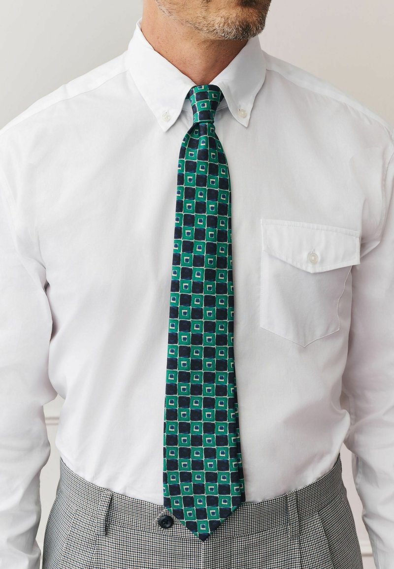 Muž v bielej košeli s gombíkmi, vzorovanou zeleno-modrou geometrickou kravatou a sivými károvanými nohavicami, zobrazený od krku po pás.