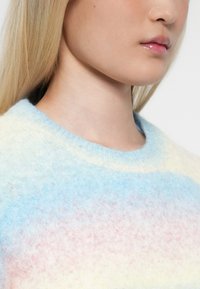Κοντινό πλάνο του κάτω μέρους ενός προσώπου και του λαιμού ενός ατόμου που φοράει ένα μαλακό, αφράτο πουλόβερ με παστέλ χρώματα σε κλίμακα από μπλε, ροζ και κρέμα.