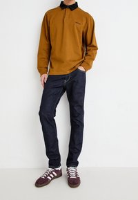 Hnědé polokošile s dlouhým rukávem a černým límečkem, kombinované s tmavými džínami a bordó teniskami s bílými pruhy.