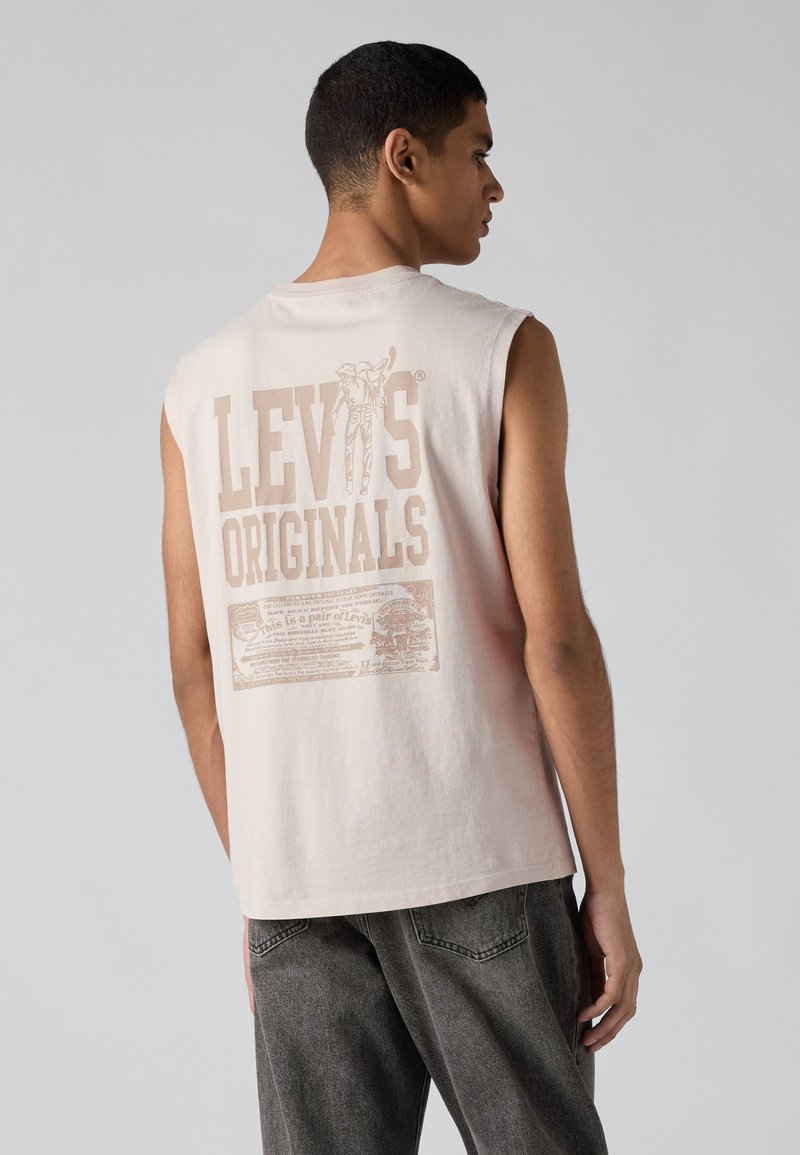 Giovane uomo che indossa una canotta beige Levi's Originals con grande logo stampato sul retro e jeans scuri larghi, di profilo.