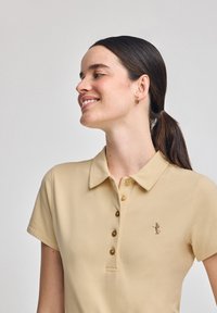 Beżowa koszulka polo o miękkiej fakturze, z kołnierzykiem, trzema guzikami i haftowanym logo na lewej piersi. Krótkie rękawy.