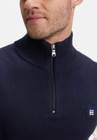 Pull-over en tricot bleu marine avec col haut et fermeture éclair quart. Dispose d'un tissu texturé et d'une petite étiquette de marque sur la poitrine gauche.