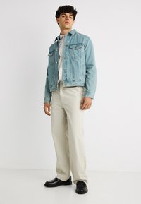 Νέος άνδρας με ανοιχτό μπλε τζιν μπουφάν, λευκό πουκάμισο, μπεζ παντελόνι και μαύρα παπούτσια, στέκεται μπροστά σε απλό λευκό φόντο.