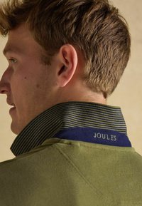 Olivgrüner Kragen mit marineblauem und gestreiftem Innenfutter, besticktes "JOULES"-Detail, strukturiertes Gewebe, Nahaufnahme des Nackens und Haares eines Mannes.