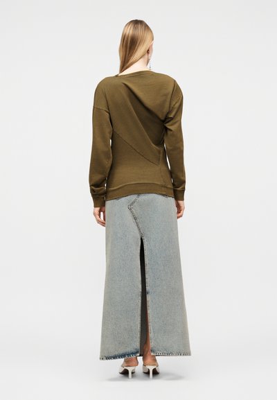 Γυναίκα που φοράει πράσινο ελιάς μακρυμάνικο τοπ και ανοιχτό μπλε τζιν μακριά φούστα με άνοιγμα πίσω, όρθια με την πλάτη στραμμένη, σε λευκές ψηλοτάκουνες γόβες.