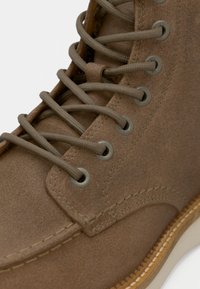Primo piano di un boot in suede marrone chiaro con lacci tondeggianti color cammello infilati attraverso occhielli metallici e dettagli di cucitura visibili lungo la suola.
