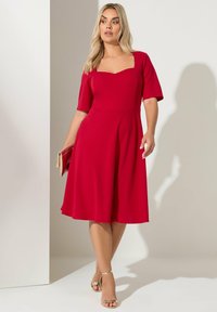 Czerwona sukienka w kształcie litery A z kwadratowym dekoltem, krótkimi rękawami, miękkiego materiału, rozkloszowanym dołem, zestawiona ze złotymi sandałami na obcasie. W ręku kopertówka.