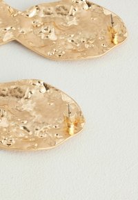 Pendientes de metal texturizado en color dorado con forma ondulada, que presentan un acabado martillado y un cierre de palanca para un ajuste seguro.