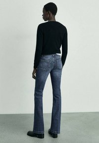 Černý svetr, rozšířené modré džíny z denimu s minimálním stárem a černé boty na podpatku. Pohled zezadu, na světlém pozadí.