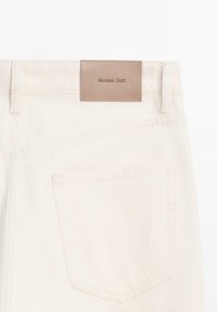 Beige denimjeans med en struktureret overflade, der har en brun læderlabel med præget "Massimo Dutti" på linningen.