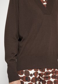 Personne portant un pull marron à col en V sur un pantalon à motifs avec de grandes formes circulaires marron et noires sur un fond blanc.
