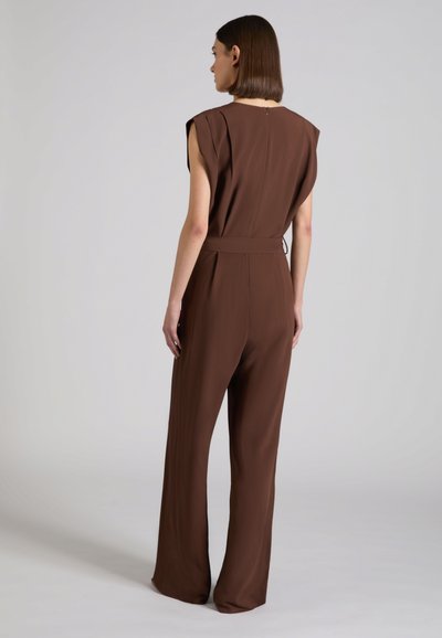 Frau mit glattem braunem Haar, die einen braunen ärmellosen Overall mit Gürtel trägt, steht mit dem Rücken zur Kamera vor einem schlichten grauen Hintergrund.