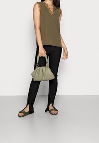 Oliviinivihreä hihaton paita, jossa on pitsikoristelu, yhdistettynä mustiin muotonsa mukaisiin housuihin, joissa on halkiot. Malli pitää kädessään vaaleanvihreää, teksturoitua käsilaukkua.