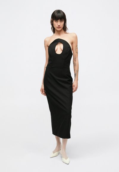 Μαύρο φόρεμα με λαιμόκοψη halter, άνοιγμα στο στήθος σε σχήμα κλειδιού, εφαρμοστή σιλουέτα και μήκος έως το γόνατο. Το μοντέλο φοράει κρεμ τριγωνικά τακούνια.