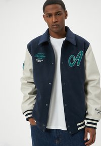 Giacca varsity blu navy con maniche grigie chiare, caratterizzata da scritte ricamate e una grande lettera "A" verde. Polsini e colletto a righe a coste.