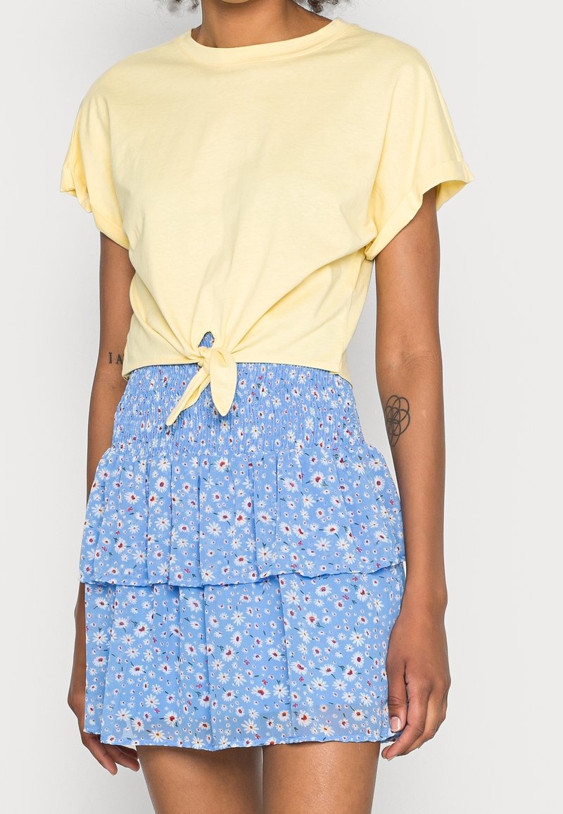 Żółta krótka koszulka z związanym przodem, połączona z niebieską spódnicą w kwiatowy wzór z falbanami i elastycznym pasem.