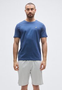 T-shirt en coton bleu à manches courtes et col rond, avec un petit logo jaune sur la poitrine, associé à un short gris clair.