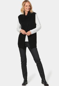 Schwarze ärmellose Strickweste mit einem Schalkragen, kombiniert mit einem weißen Langarmshirt und schwarzen eng anliegenden Hosen, getragen mit Stiefeletten.