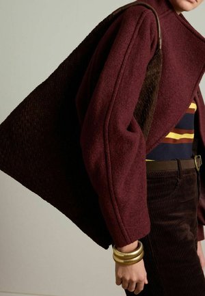 Bordeaux wollen jas met gestructureerde schouders gecombineerd met een donker geweven draagtas. Het model draagt een gestreept topje en gouden armbanden.