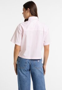Camicia a maniche corte, a righe rosa chiaro con colletto classico. Indossata con jeans a gamba larga blu con un accento a forma di triangolo sulla tasca posteriore.