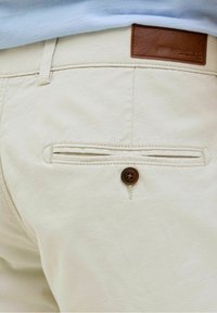 Pantalon beige avec poche passepoilée boutonnée à l'arrière et écusson en cuir marron sur la ceinture, porté avec une chemise bleu clair.