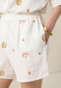 Shorts en coton blancs ornés de broderies en forme de coquillage dans des tons dorés et roses, avec une taille élastique, des poches latérales et des détails décoratifs au niveau de l'ourlet.