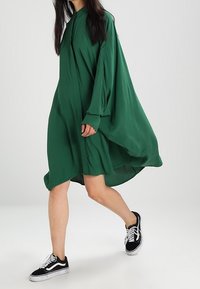 Vestito verde oversize realizzato in tessuto leggero, con colletto alto e maniche lunghe, abbinato a sneakers nere.