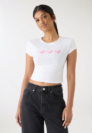 T-shirt bianca corta a maniche corte con tre cuori rosa chiaro disegnati sul davanti. Abbinata a jeans neri a vita alta.