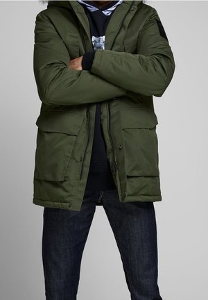 Personne portant une veste d'hiver verte avec de grandes poches avant, un sweat à capuche noir et un jean bleu foncé, debout les bras croisés.