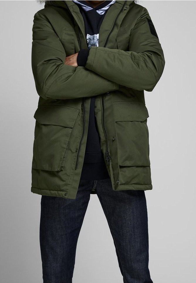 Personne portant une veste d'hiver verte avec de grandes poches avant, un sweat à capuche noir et un jean bleu foncé, debout les bras croisés.