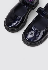 Zapatos Mary Jane de charol azul marino con patrones decorativos perforados y suelas gruesas de goma negra, descansando sobre una superficie gris claro.