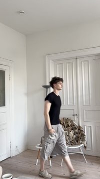 Nuori mies mustassa t-paidassa ja harmaissa shortseissa kävelee valkoisessa huoneessa, jossa on tuoli, jonka päällä on turkkipeitto, sekä lattiavalaisin.