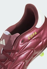 Botas de fútbol burdeos con parte superior texturizada y brillante, cuello blanco en contraste y detalle "COPA" en amarillo con cordones negros.