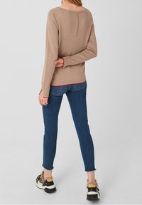 Kobieta w beżowej bluzce z długim rękawem i guzikami z tyłu, niebieskich wąskich dżinsach z postrzępionymi brzegami oraz sneakersach z futrzanym wykończeniem, stojąca na tle jednolitym.