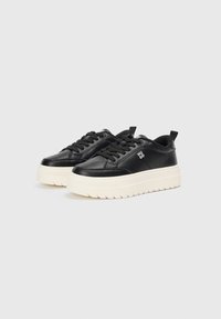 Zapatillas de cuero negro con gruesa suela de goma blanca, puntera redondeada, diseño de cordones y logo visible en el lateral. Detalles texturizados en la suela.