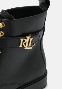 Botín negro de cuero hasta el tobillo con acrílico de metal dorado, puntera redonda, cordones en la parte delantera y un diseño elegante. Textura suave con detalles de costura.