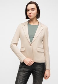 Jeune femme aux cheveux lisses et foncés portant un blazer beige sur un haut vert et un pantalon noir texturé, debout devant un fond clair uni.