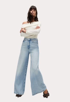 Jeans a vita alta di denim blu chiaro a gamba larga, abbinati a un top bianco scollato sulle spalle e scarpe con tacco nere.