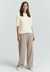 Donna con lunghi capelli castani che indossa un maglione corto a maniche corte bianco con texture, pantaloni beige a gamba larga e sneaker bianche, in piedi su uno sfondo semplice.